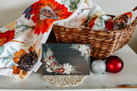 Decorative basket with floral blanket, ornaments, and a 'Seasons Greetings' card on a wooden surface.
