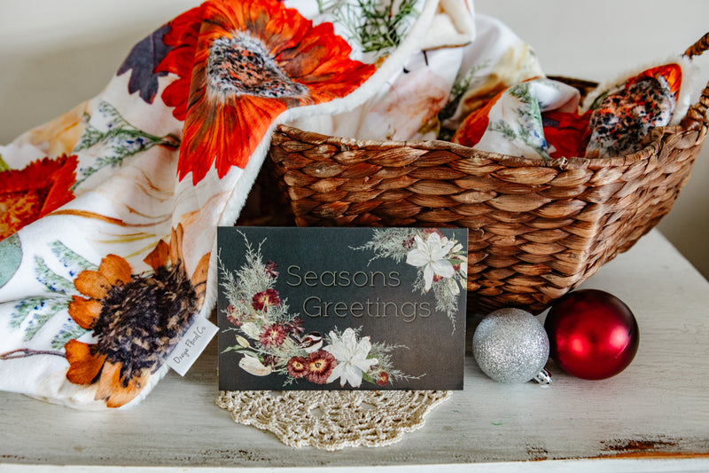 Decorative basket with floral blanket, ornaments, and a 'Seasons Greetings' card on a wooden surface.