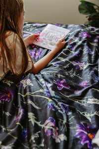 Person sitting on a floral-patterned bedspread, reading a book.