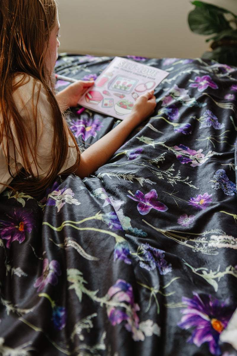 Person sitting on a floral-patterned bedspread, reading a book.