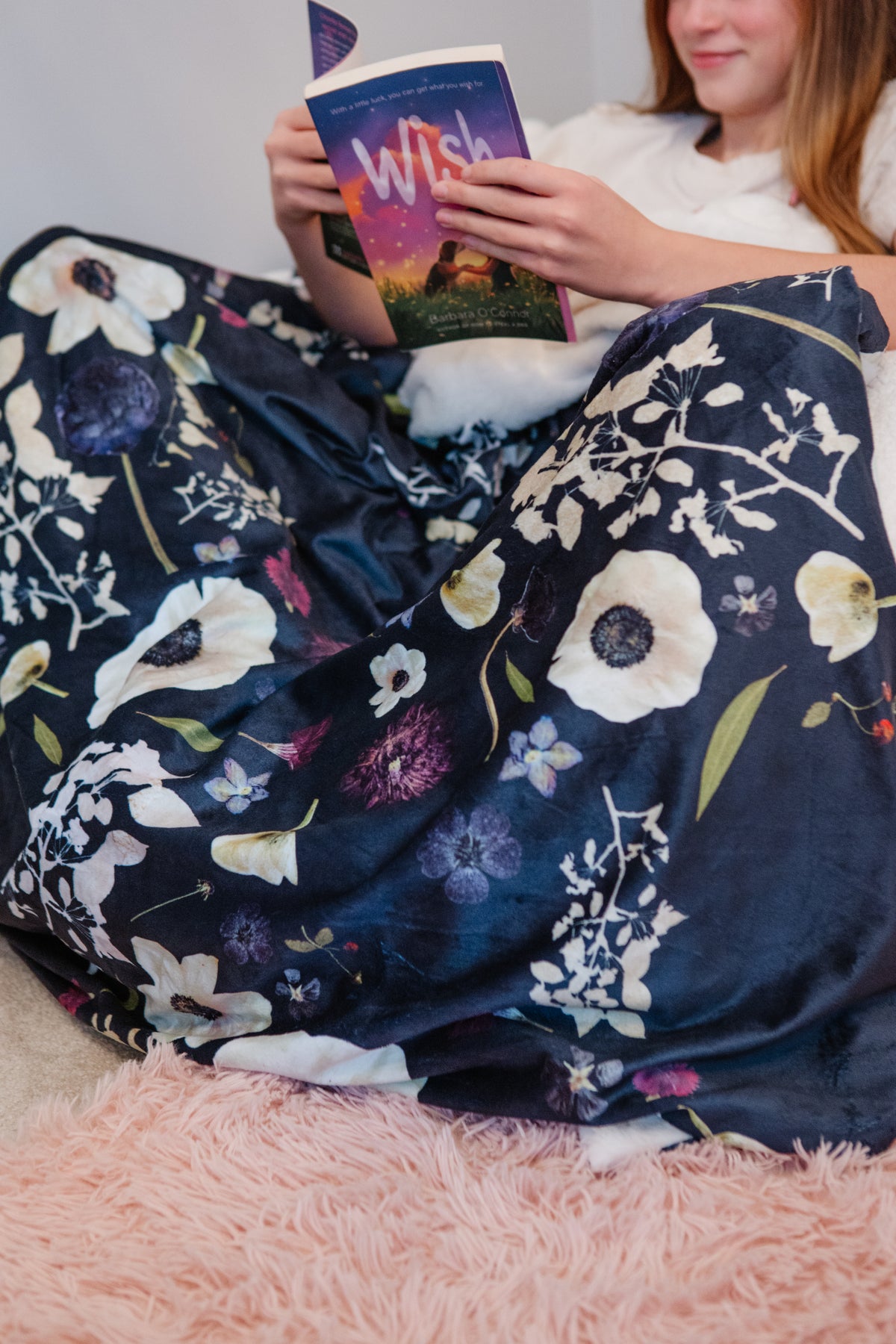 Person reading a book under a floral blanket on a pink surface