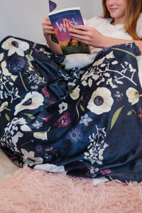 Person reading a book under a floral blanket on a pink surface