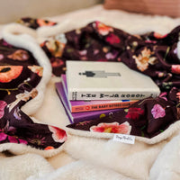 A cozy floral throw blanket with a white fluffy backing, styled with a stack of books on top. The blanket features a dark purple floral pattern with blush pink and white flowers, and a small white tag reading 'Oxeye Floral Co' is visible.