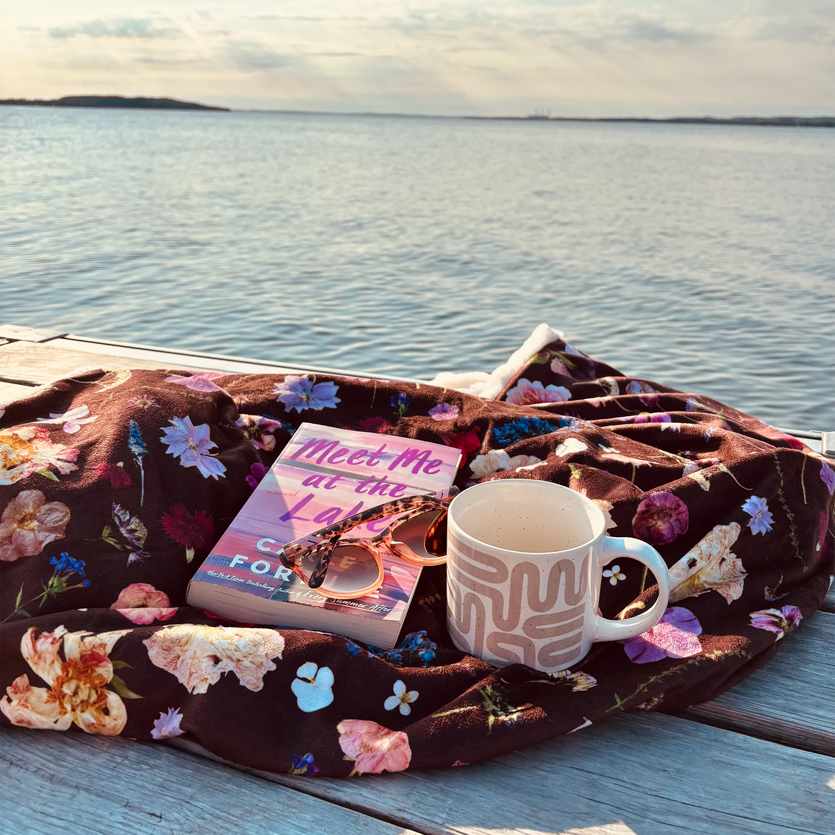 A cozy scene on a wooden dock with a body of water in the background. A floral blanket in dark burgundy with blush pink, purple, and white flowers is spread out, holding a book, a pair of sunglasses, and a white ceramic mug.