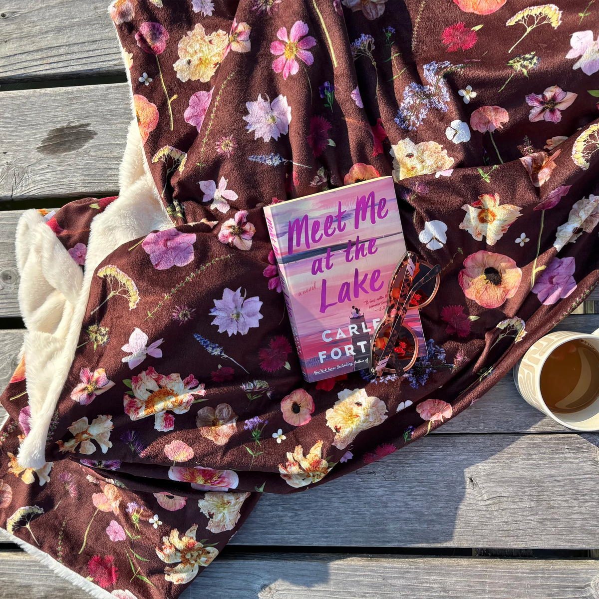 A cozy floral blanket in burgundy with pink, purple, and white pressed flowers is spread out on weathered gray wooden decking. A pink book, a pair of sunglasses, and a mug of tea are placed on top of the blanket.
