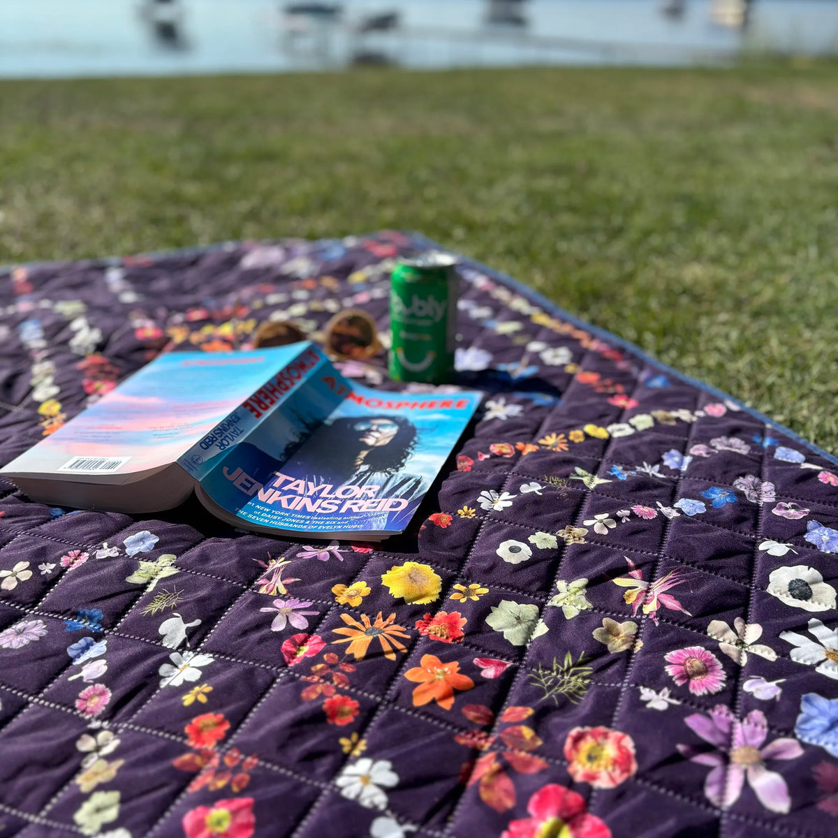 Flower-patterned blanket with books and a can on grass by water