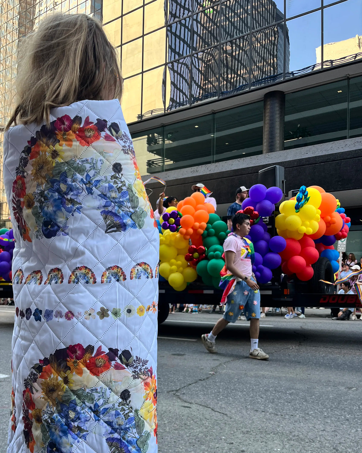 Person wrapped in a colourful quilt with rainbow patterns, walking past a float with balloons in an urban setting.