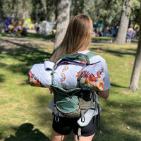Person with a floral-patterned blanket rolled up and attached to their backpack in a park setting