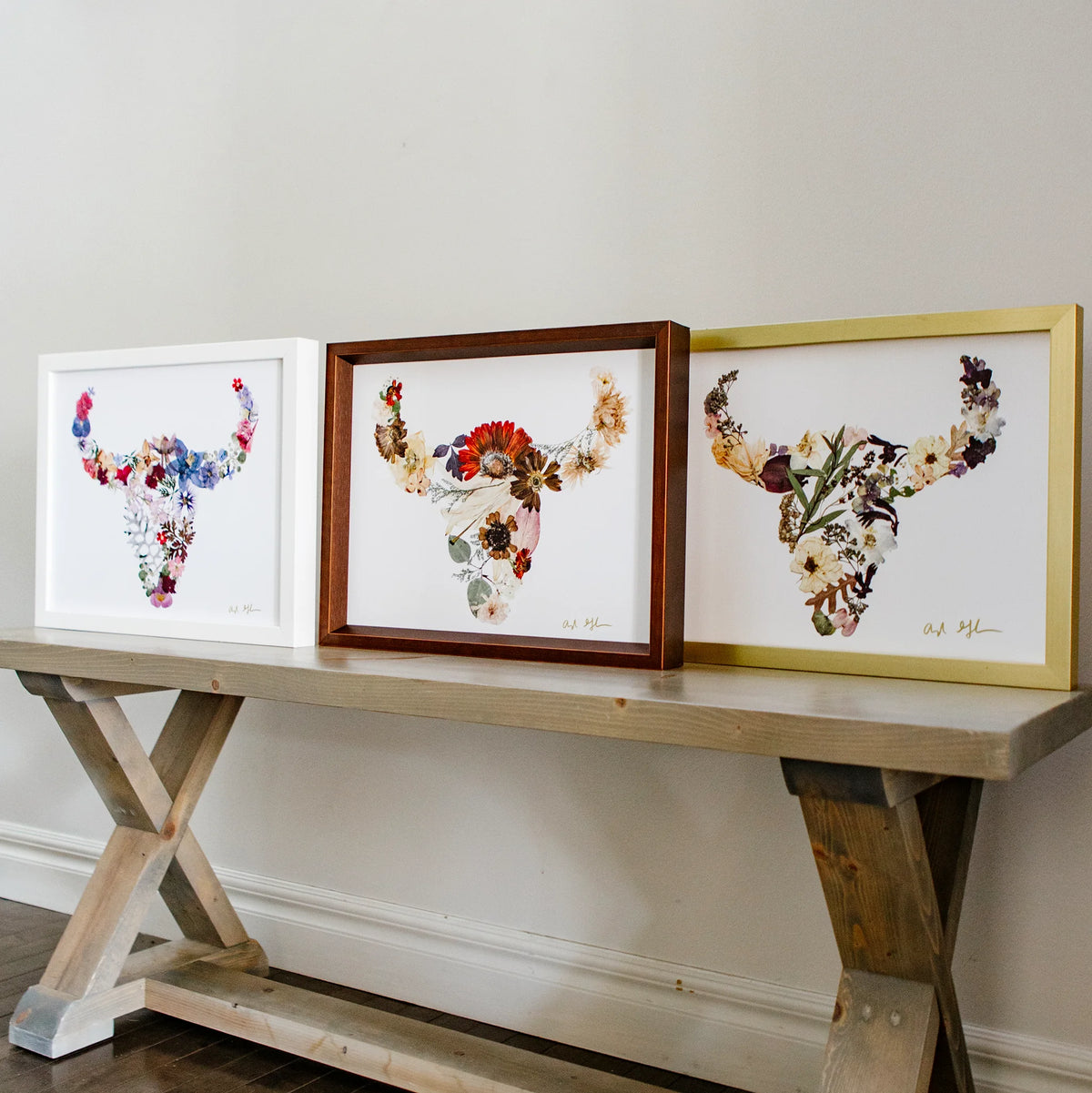 Three framed floral buffalo art pieces on a wooden console table against a white wall.