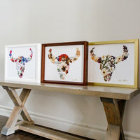 Three framed floral buffalo art pieces on a wooden console table against a white wall.