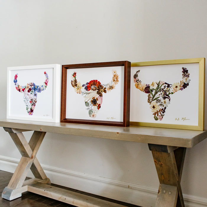 Three framed floral buffalo art pieces on a wooden console table against a white wall.