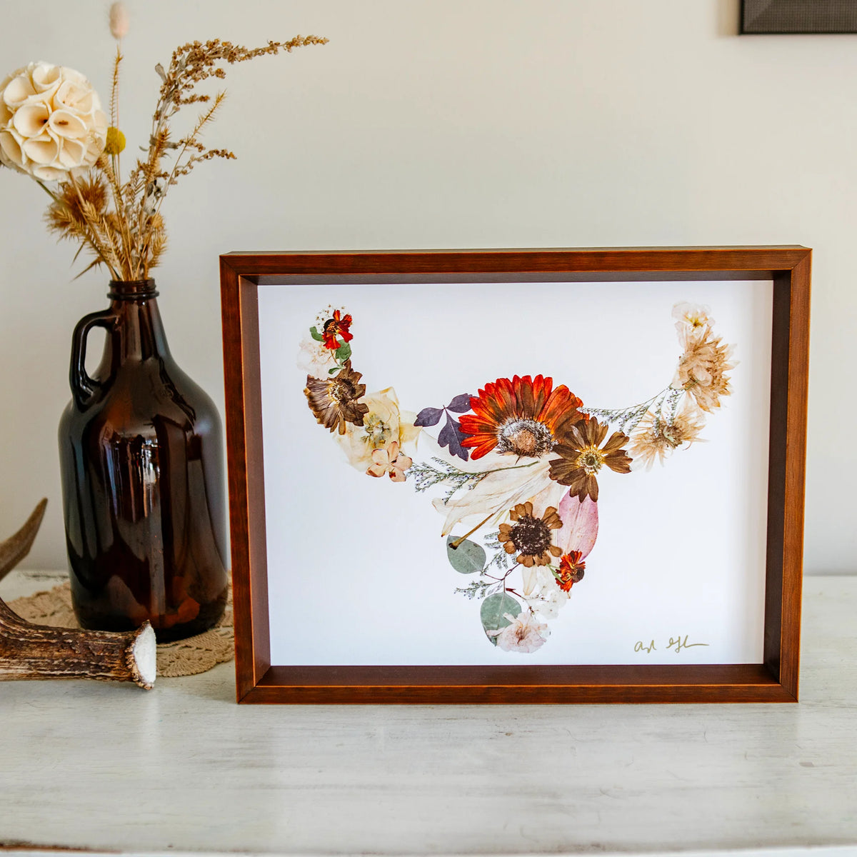 Framed floral buffalo artwork on a table with a vase and antlers in the background