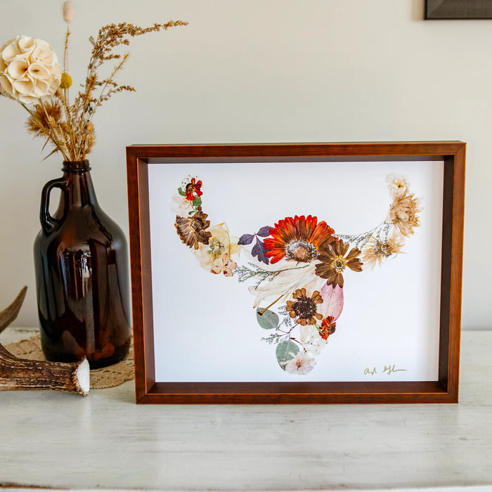 Framed floral buffalo artwork on a table with a vase and antlers in the background