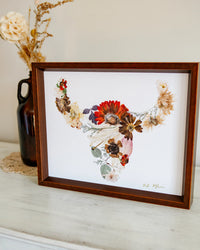 Framed floral artwork on a white surface with a vase of flowers in the background.