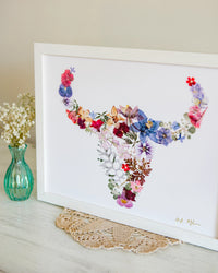 Framed artwork of floral buffalo skull on a table with a vase and doily.
