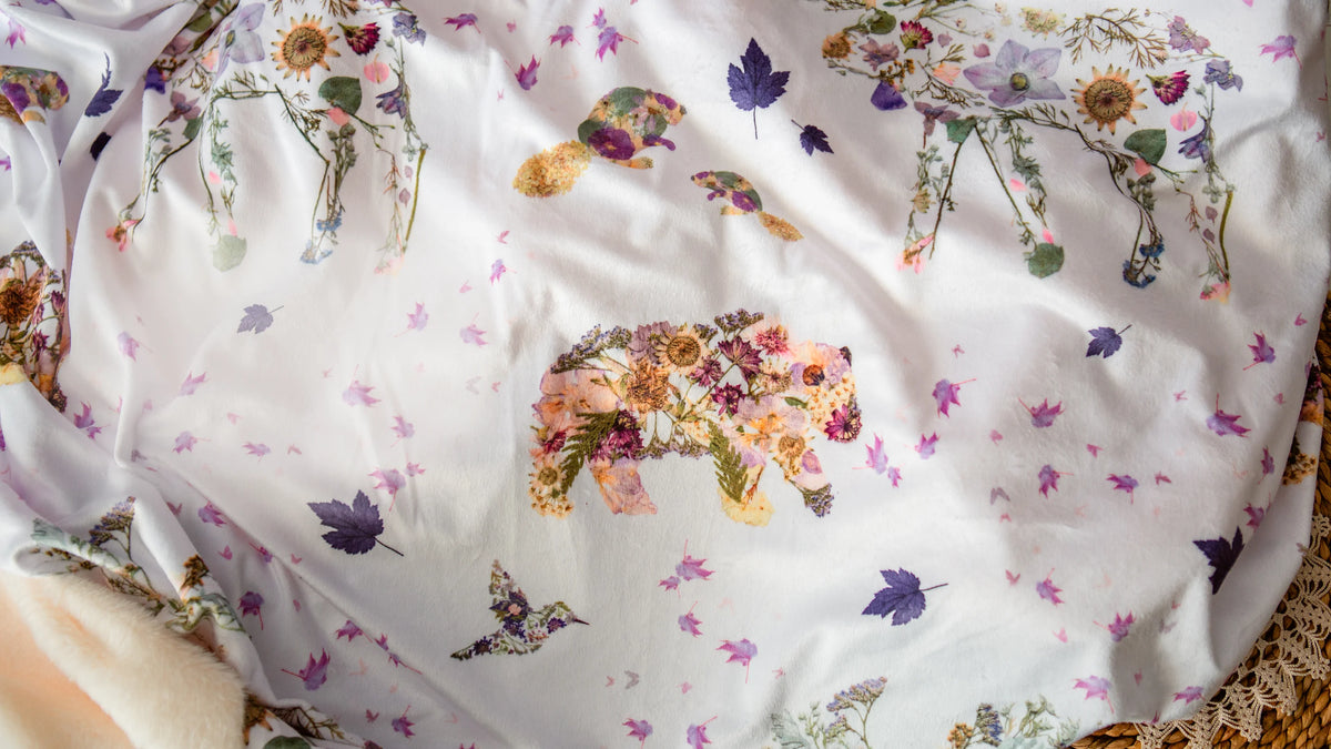 White blanket with a bear, hummingbird and other Canadian animals made from pressed flowers, scattered with maple leaves