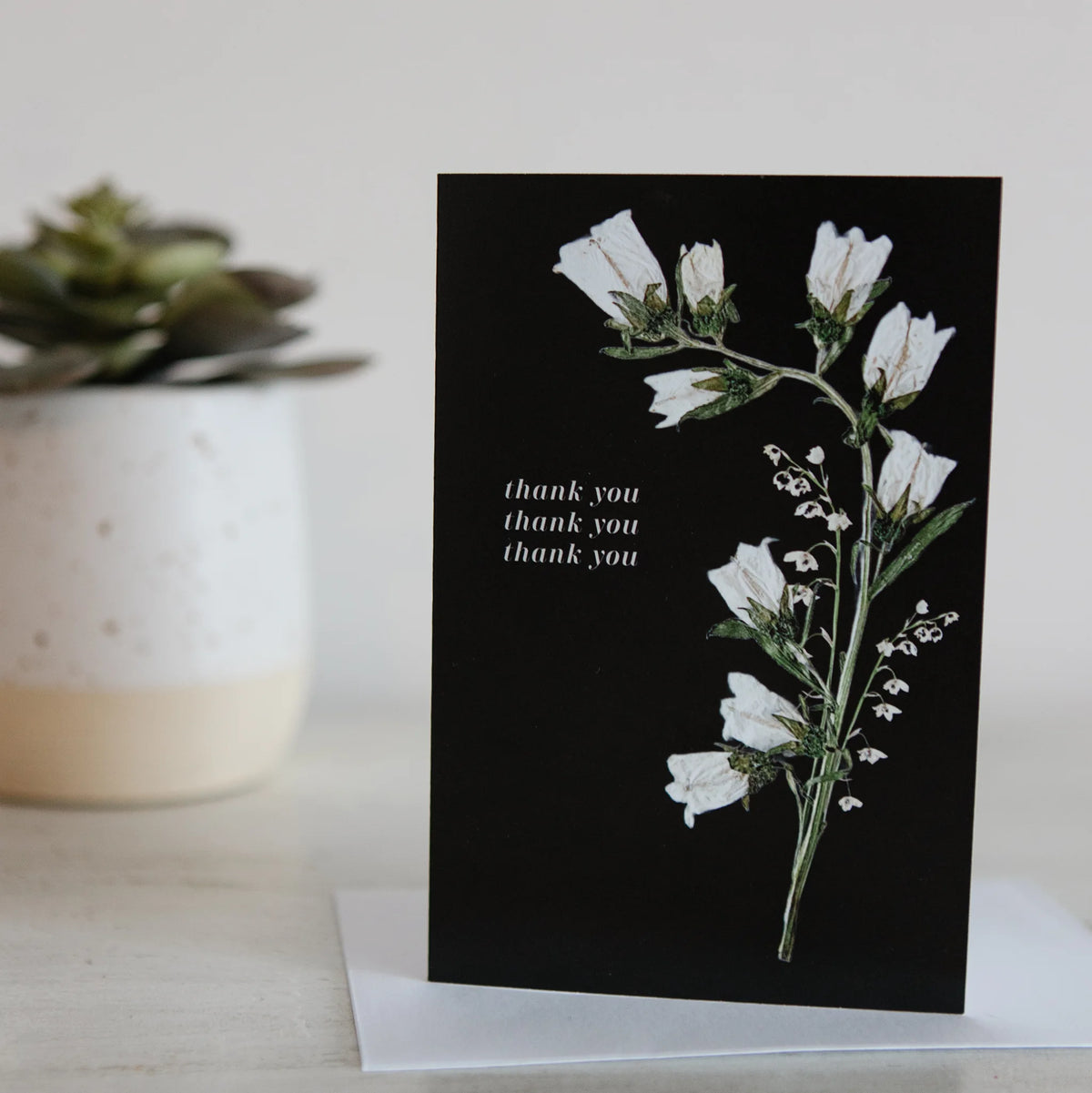 Greeting card with white floral design and 'thank you' text on a black background, placed on a light surface with a plant in the background.
