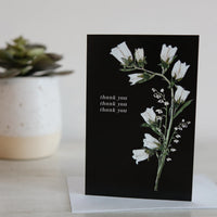 Greeting card with white floral design and 'thank you' text on a black background, placed on a light surface with a plant in the background.