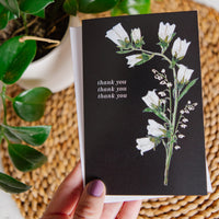 Card with white floral design and 'thank you' text held by a hand on a woven surface.