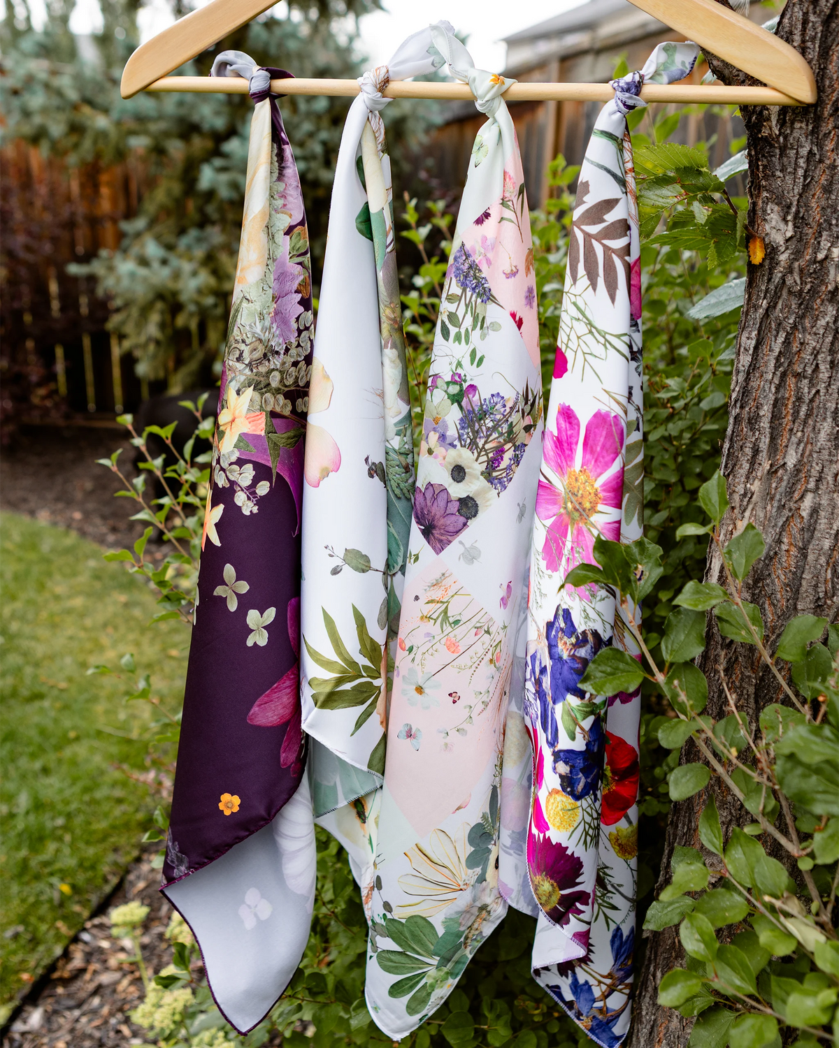Four floral silk squares hanging from a wooden dowel in a garden setting
