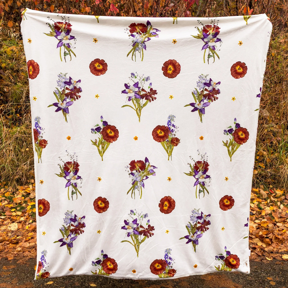Cream-white blanket with flower cluster design being held up in an autumn setting outdoors