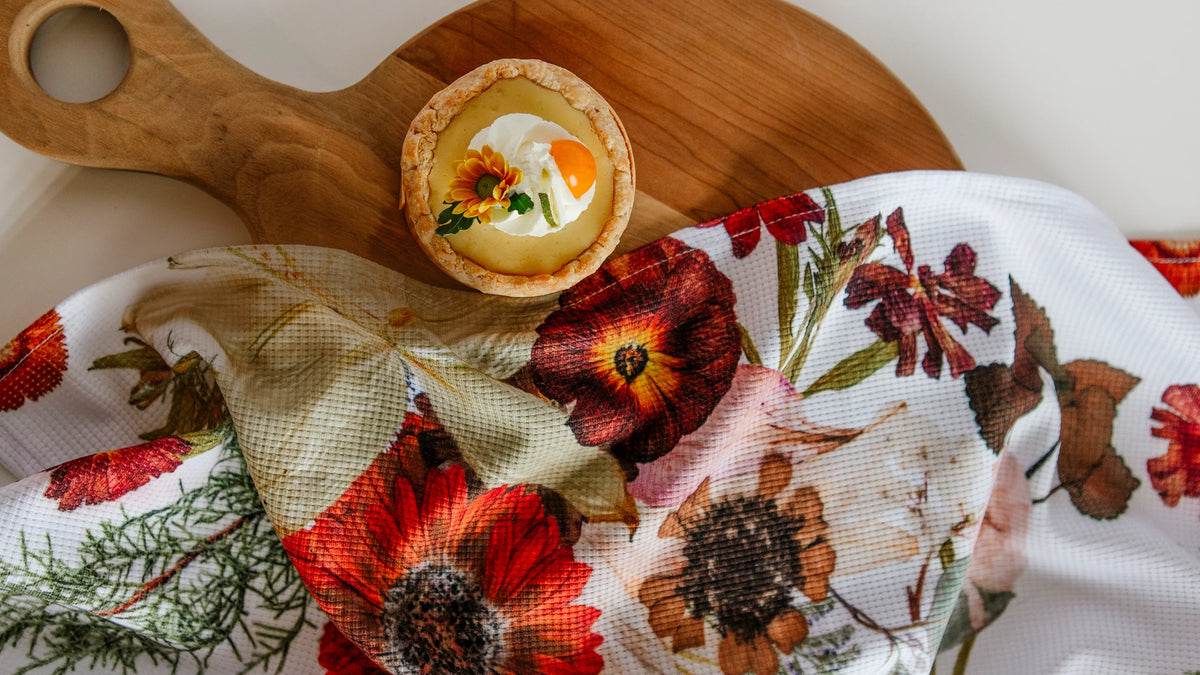 Floral waffle-weave tea towel with red and orange pressed flower print, beside a tart on a wooden cutting board