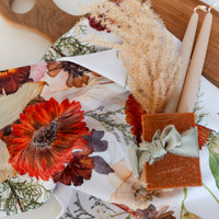 Decorative table setting gift set with a floral tea towel, candles, and a soap bar on a wooden surface.