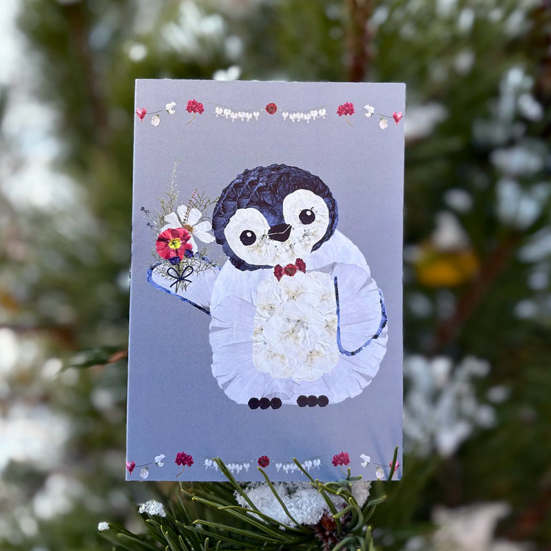 Card with a penguin holding flowers on a gray background, surrounded by greenery.