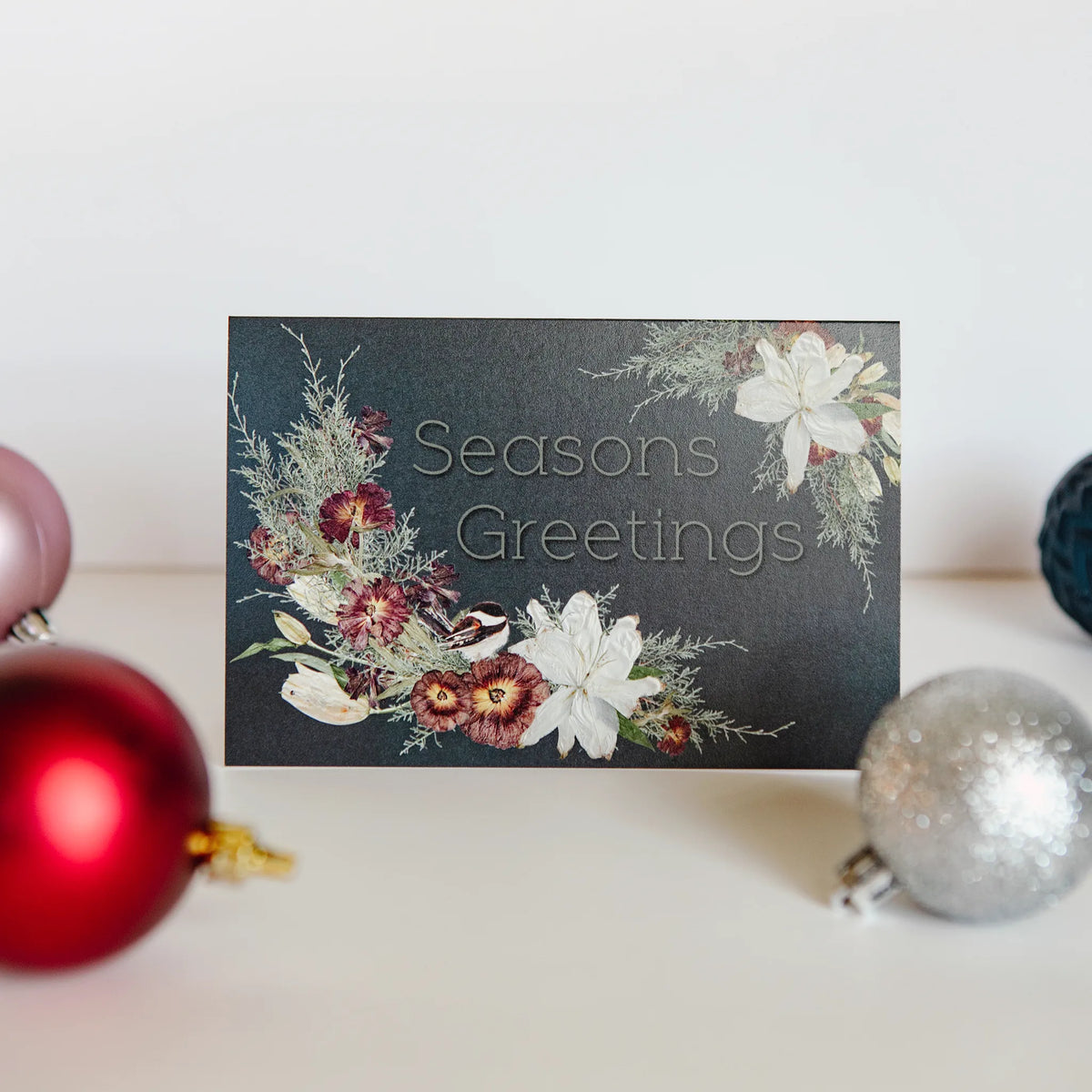 Christmas card with floral design and 'Seasons Greetings' text, surrounded by ornaments on a light background.