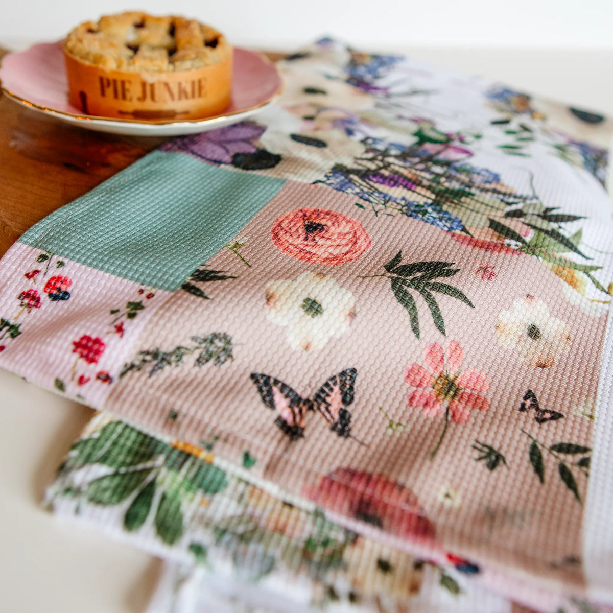 Floral and butterfly patterned tea towel with a pie on a pink plate in the background