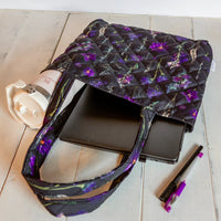 Black and purple pressed flower tote bag with laptop and water bottle on a neutral wood background